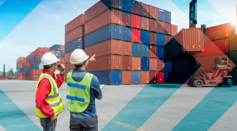 Two workers wearing helmets and safety vests are standing in front of stacked cargo containers, overseeing the managed transportation process. One points towards a container being lifted by a crane in the busy shipping yard under a clear blue sky.