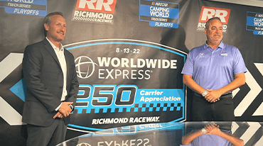 Two men stand in front of a promotional backdrop for the Worldwide Express 250 Carrier Appreciation event at Richmond Raceway. The backdrop features logos and event details. Both men are smiling and wearing business attire.