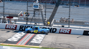 A blue race truck with the number 42 speeds past the checkered finish line at a racetrack with RICHMOND painted on the asphalt. Empty bleachers and a few spectators are visible in the background.