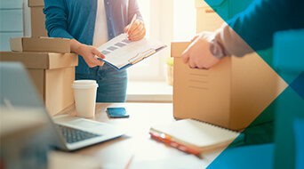 In a well-lit room, two people manage boxes with precision. One holds a clipboard, the other a cardboard box. A laptop, coffee cup, and notebook rest on the table, painting a picture of an efficient workspace—perhaps inspired by practices from the best 3PL solutions.