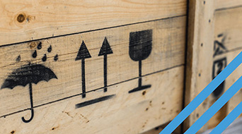 Symbols on a wooden crate illustrate handling instructions: a rain-protected umbrella, two upward arrows, and a fragile wine glass. These icons are crucial for understanding BOL Shipping guidelines. Blue diagonal stripes mark the right edge of the image.