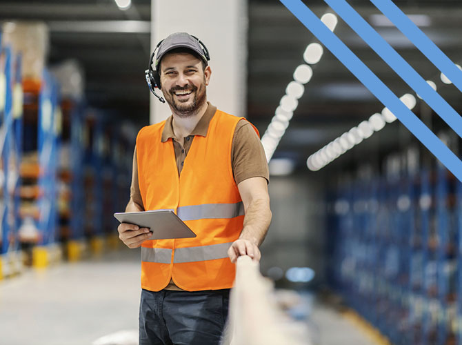 A man in a safety vest and cap stands in a 3PL warehousing facility. He smiles while holding a tablet, headset on, amidst tall shelves and bright overhead lighting.