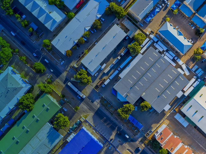 Aerial view of an industrial area with large rectangular buildings, likely 3PL warehousing facilities, and parking spaces. The structures have roof colors in white, blue, and green. Trees and roads weave between the buildings, creating a grid-like pattern.