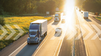 Two semi-trucks from a freight carrier and two cars drive on a highway at sunrise. The brilliant glare of sunlight illuminates the way, with the road lined by grass and trees. White arrows are digitally superimposed, pointing right, suggesting motion.