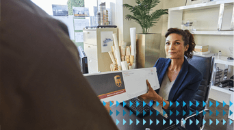 A woman in a blue blazer sits at a desk and hands a package to a person off-frame. The room has shelves with small decorative items and a large plant. A pattern of blue arrows overlays the bottom corner of the image.