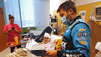 A masked man in a blue WWEX NASCAR racing suit signs an autograph for a young patient in a hospital bed. A woman in pink stands nearby, holding a phone, capturing the heartfelt moment. The room, brightened by natural light from the large window, is filled with medical equipment.