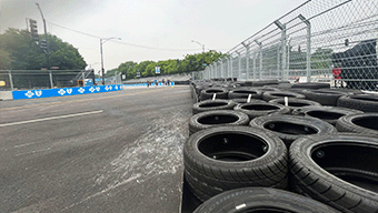 The street race track features a row of stacked tires on the right, bordered by fencing. Trees and streetlights line the backdrop against a cloudy sky, reminiscent of the thrilling atmosphere youd find at a WWEX NASCAR event in Chicago.