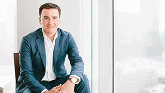WWEX executive Joel Clum sits in a blue suit and white shirt by a large window with a sweeping cityscape view. The bright, modern room complements his confident smile as he gazes towards the camera, embodying professionalism and approachability.