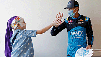 A child in a printed headscarf and pajamas high-fives a masked racecar driver from WWEX NASCAR, dressed in a blue and black racing suit. The driver holds papers as they stand in a brightly lit room, capturing the spirit of Richmonds racing excitement.