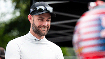 A man in a white racing suit and black cap beams outdoors, embodying the spirit of WWEX NASCAR. Sunglasses rest on his cap above his beard. In the foreground, another person sports a helmet adorned with an American flag pattern, reminiscent of iconic races with Shane van Gisbergen.