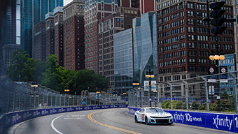 A race car speeds through the urban circuit of the WWEX NASCAR Chicago event, surrounded by towering buildings. It is daytime, and the road is lined with Xfinity ads and protective barriers, while lush green trees contrast with both modern and historic architecture in the background.