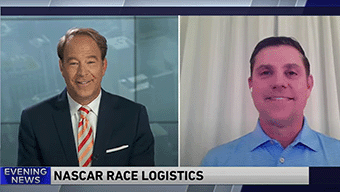 A news broadcast shows two men on a split screen discussing NASCAR race logistics. The left side features a suit-clad man labeled Evening News, while the right side shows another in a blue shirt with earbuds, focusing on the impact of WWEX sponsorship at the Chicago event.
