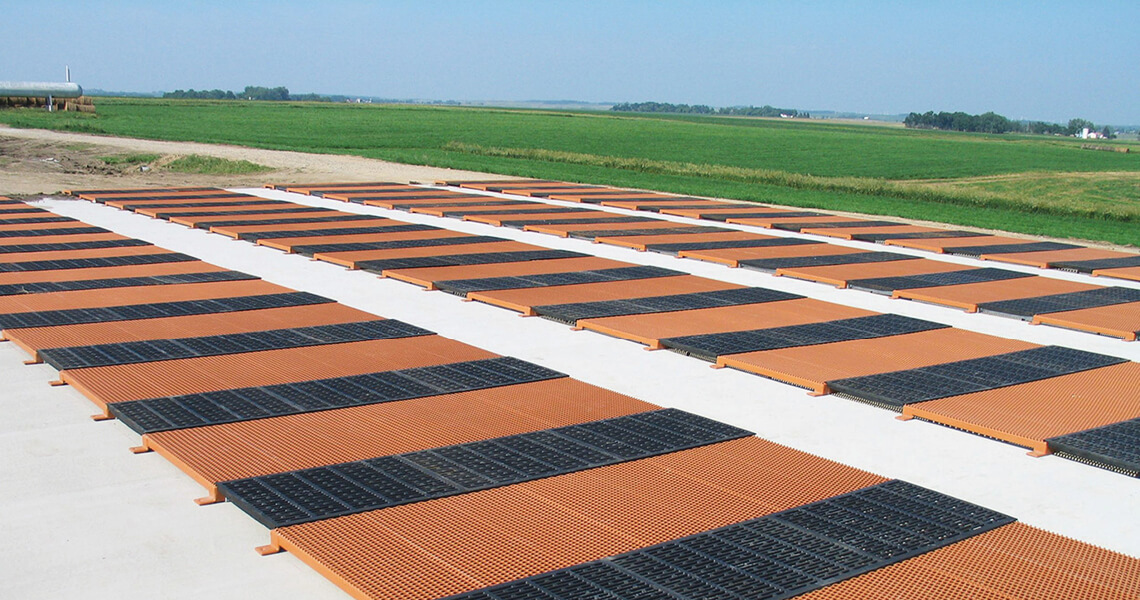 A large outdoor area with multiple rectangular solar panels arranged on a flat surface. The panels are aligned in rows and columns, with alternating sections of black and orange grids. A vast green field stretches into the distance under a clear blue sky.