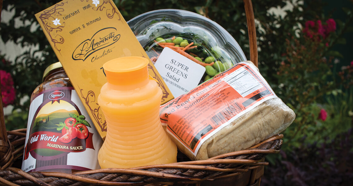 A wicker basket containing a bottle of Old World marinara sauce, a plastic jug of orange juice, a packet of chocolate, a container labeled Super Greens Salad, and packaged cheese, set against a garden backdrop.