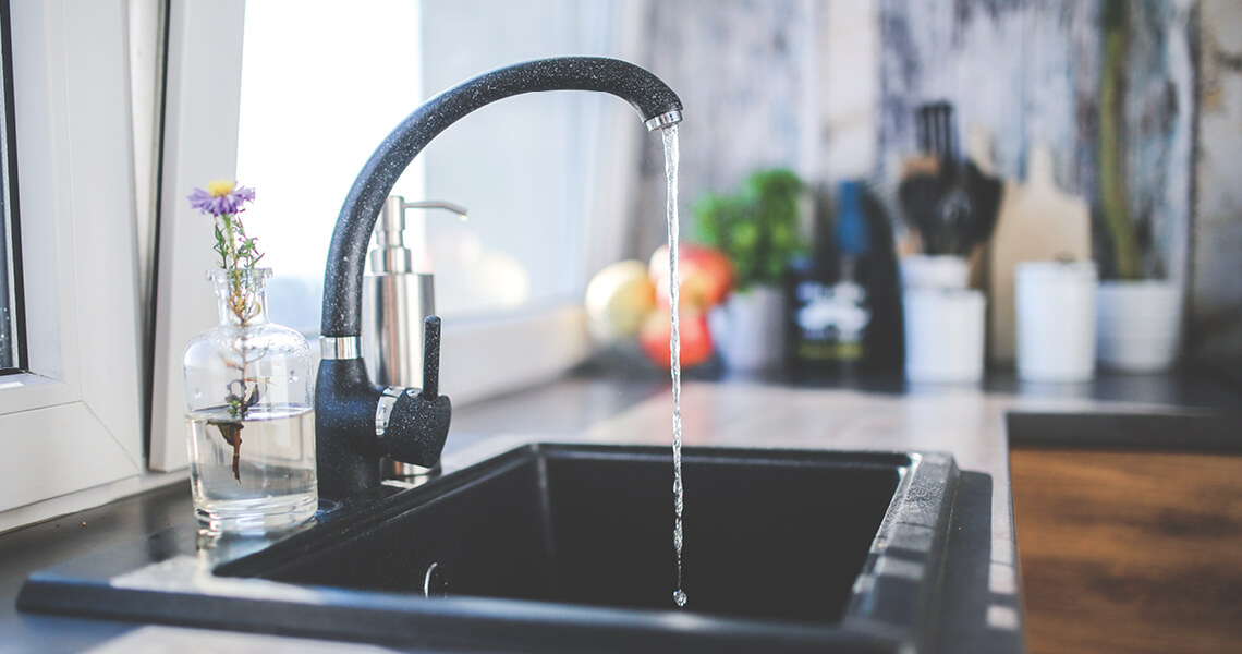 A kitchen sink with a black faucet is running water. A small vase with flowers and a soap dispenser are on the countertop. The background is slightly blurred, showing kitchen items and natural light coming through a window.