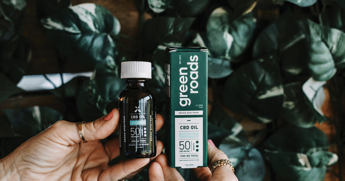 Two hands holding a bottle and box of Green Roads CBD Oil against a leafy background. The bottle reads 50 mg and Broad Spectrum. The box is green with white text, showcasing the same product details.
