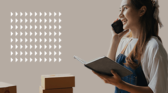 A woman with long hair stands smiling, holding a phone to her ear and a notebook in her other hand. She wears a white shirt and denim overalls. Several cardboard boxes are stacked nearby. The background is beige with a pattern of white arrows.