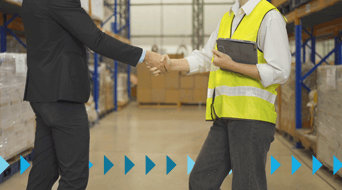 A person in a business suit and a person in a yellow safety vest shake hands in a bustling warehouse, where shelves brim with boxes. Blue arrows guide the way for efficient freight shipping services in the background, ensuring seamless logistics.