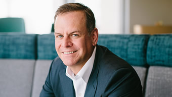 A smiling man in a business suit sits on a gray and teal cushioned bench, embodying the professional spirit of Worldwide Express as they unveil WWEX Group. With short dark hair and a crisp white shirt, he relaxes in the brightly lit room, the softly blurred background setting a composed scene.