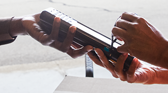Close-up of two people exchanging an elongated handheld device with a strap, reminiscent of a UPS Signature Required delivery. The background is blurred, emphasizing their hands and the device.