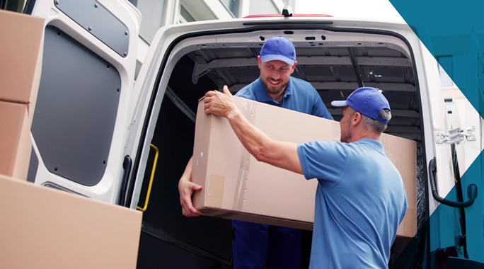 Two men in blue uniforms are efficiently unloading large cardboard boxes from a white delivery van, showcasing their white glove freight service. One man is inside the van, carefully handing a box to the other standing outside. The scene exudes professionalism at a delivery site.