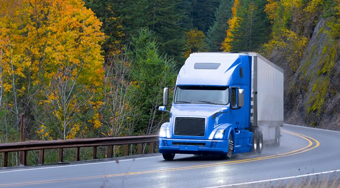 A blue semi-truck navigates a wet, winding road, reflecting the current state of the shipping industry. Its flanked by a forest where autumns orange and yellow leaves contrast with evergreen trees, while slick surfaces from recent rain add to the journeys challenges.