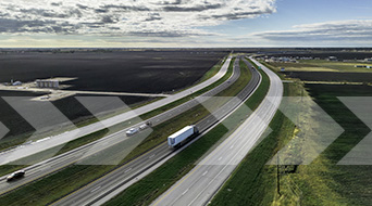 Aerial view of a four-lane highway stretching into the distance. A truck and several cars travel along the road, surrounded by vast fields under a partly cloudy sky. The landscape is open and flat, with a horizon line in the distance.