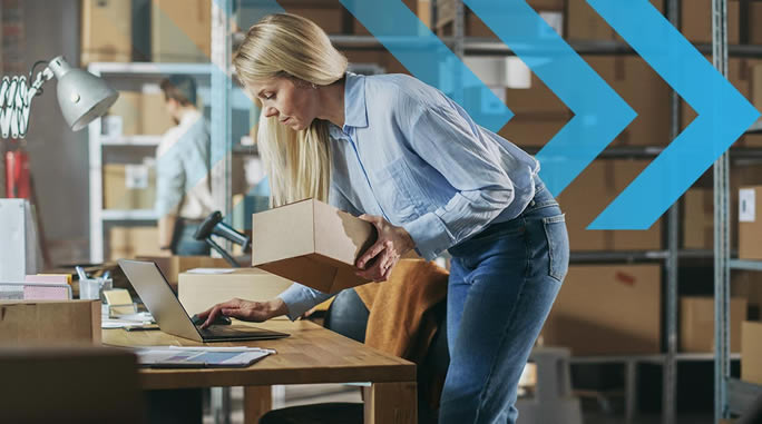 A woman with long blonde hair, wearing a light blue shirt and jeans, is holding a cardboard box and working on a laptop at a desk. The background has shelves with more boxes and a person in white standing in the distance. Blue arrows overlay the image.