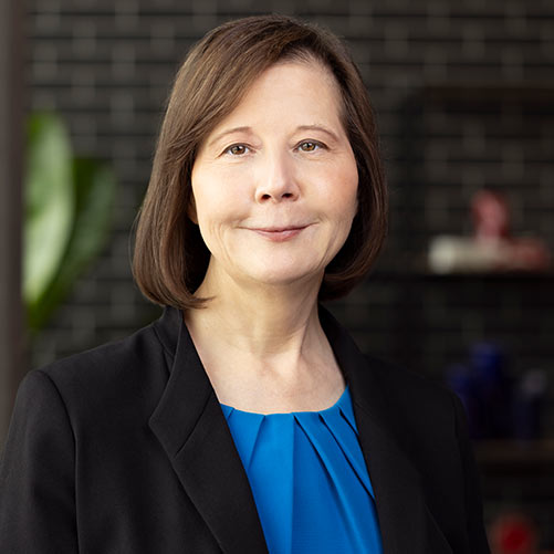 A woman with shoulder-length brown hair is smiling slightly. She is wearing a blue blouse and a black blazer. The background is blurred with dark-colored bricks and some indistinct objects, possibly decor items or plants.