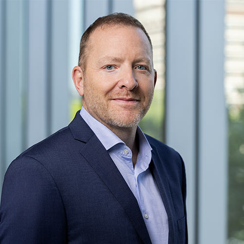 Man in a suit with a light blue shirt smiles slightly. He stands in front of large windows with blurred cityscape visible through them. The background is softly lit, creating a professional atmosphere.