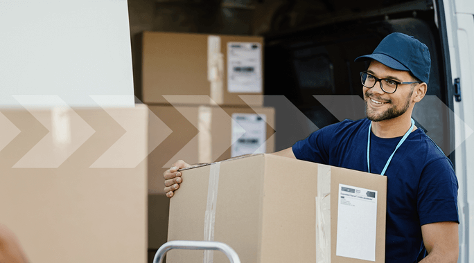 A delivery person in a blue shirt and cap is smiling while unloading a large cardboard box from a van. Several other boxes are visible inside the van. The person wears glasses and appears to be working efficiently.