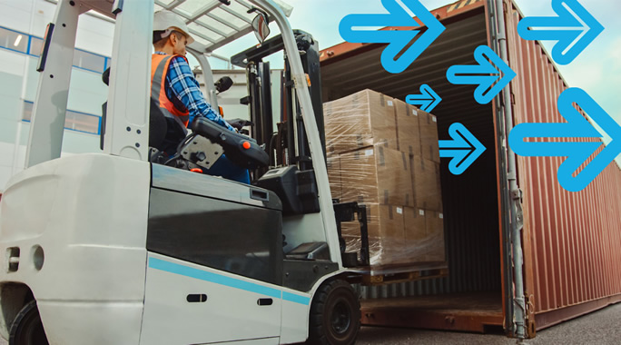 A person operates a forklift, loading wrapped pallets into a shipping container. Blue arrows overlay the image, pointing into the container, indicating movement or direction. The scene takes place outdoors near a building.