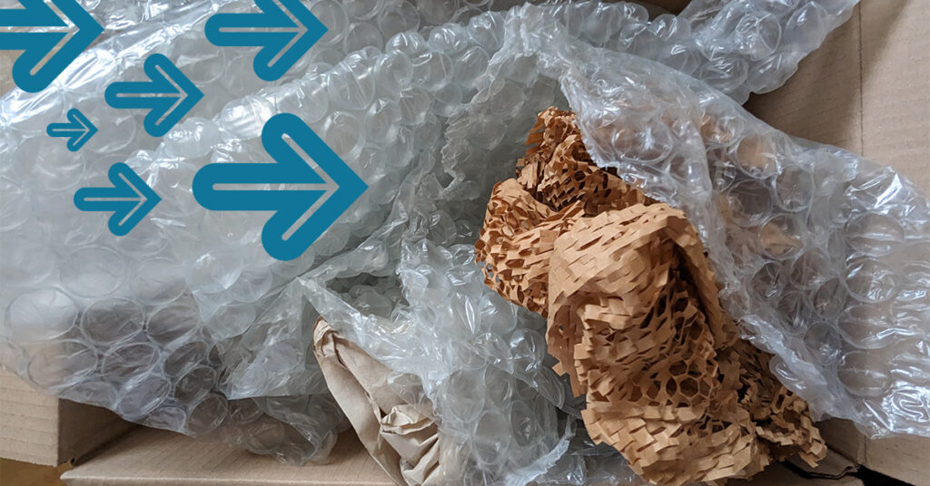 A cardboard box containing crumpled brown paper and clear bubble wrap. Blue arrow graphics are overlaid on the image, directing attention to the packaging materials.