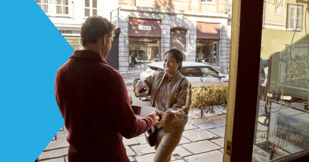A delivery person in a uniform hands a package to a person in a maroon sweater at a doorway. Theyre smiling, and a street with parked cars and storefronts is visible in the background. A blue triangle decorates the left side of the image.