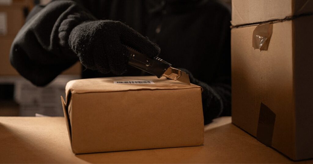 A person wearing black gloves uses a box cutter to open a sealed brown package on a dimly lit table, hinting at a storage area. Several boxes are stacked nearby, evoking an atmosphere of intrigue often associated with cargo theft.