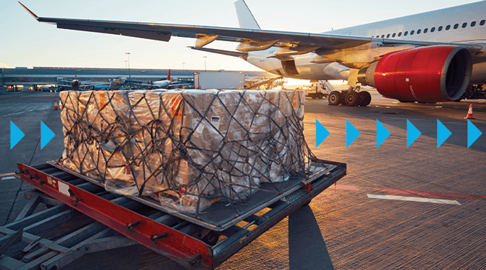 Cargo loaded on a dolly is ready for transportation in front of an airplane on a tarmac at sunset. Blue arrows indicate movement toward the aircraft.