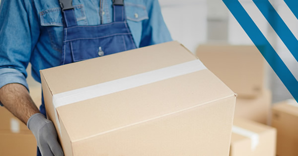A person wearing a blue shirt and overalls carries a large cardboard box sealed with white tape. Gloved hands grip the box, with more boxes and a blue diagonal stripe pattern in the background.