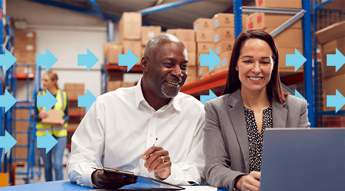Two people, one holding a tablet, smile while looking at a laptop in a warehouse with boxes. A worker in a safety vest is in the background. Blue arrows are overlaid, pointing right, suggesting movement or progress.
