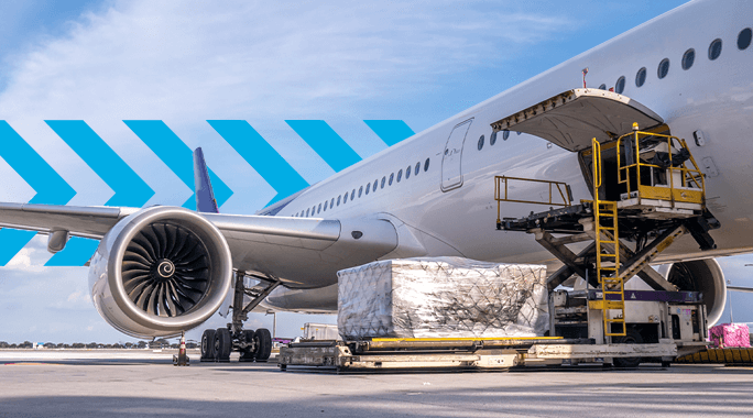 An airplane side view on an airport tarmac with an open cargo door. A cargo loading vehicle is positioned next to the plane, loading wrapped pallets onto the aircraft. The sky is clear with some clouds.
