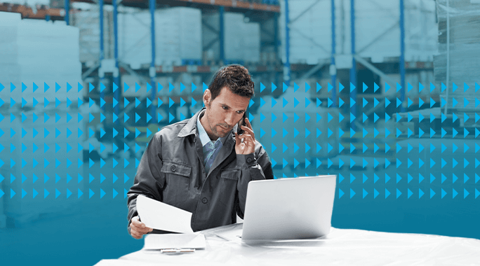 A man in a work jacket talks on the phone while looking at documents and a laptop in a warehouse with shelves and large pallets in the background.