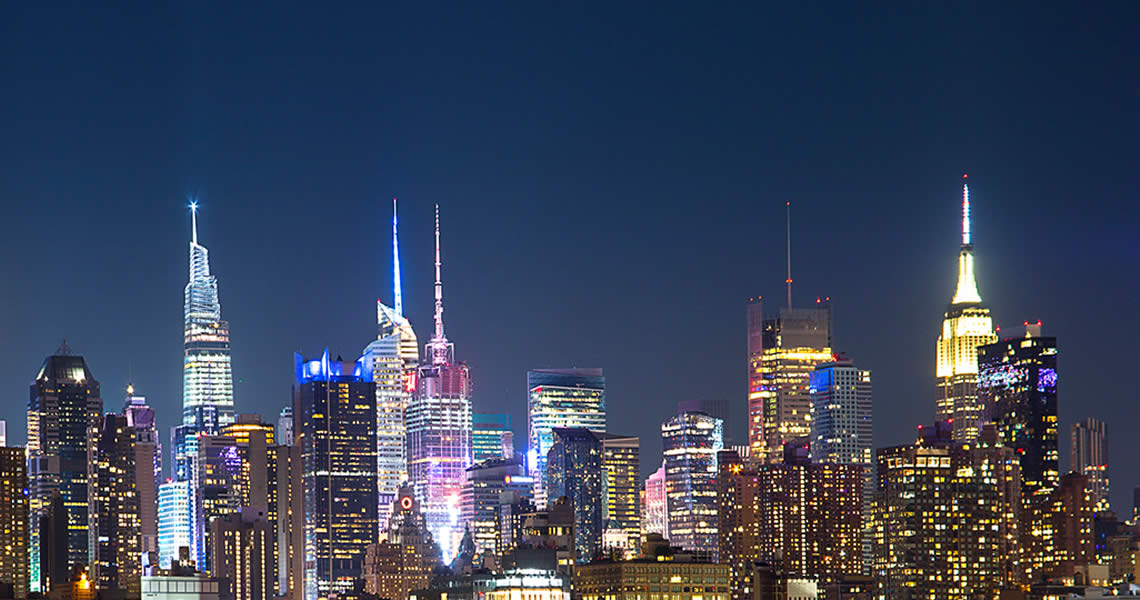 A nighttime view of a city skyline with brightly lit skyscrapers against a dark blue sky, including several tall buildings with illuminated spires and colorful lights.