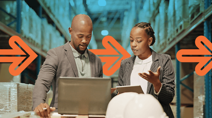 Two people in business attire discuss work in a warehouse, looking at a laptop and tablet. Shelves with boxes line the background, and large orange arrows are overlaid on the image, pointing to the right.