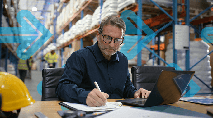 A man wearing glasses works at a desk with a laptop and papers in a warehouse, with storage shelves and workers visible in the background. Blue arrow graphics overlay the image.